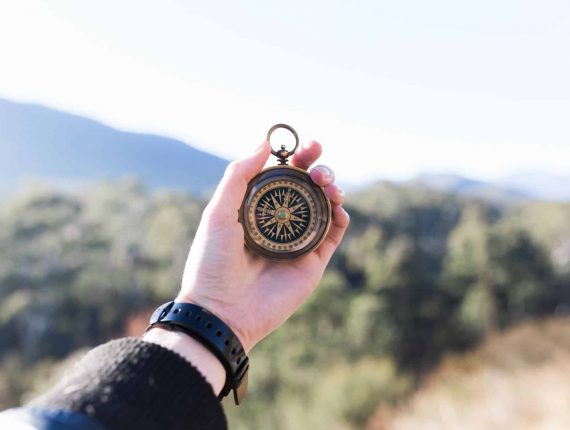 Ein ausgestreckter linker Arm mit einem Kompass in der Hand, eine schwarze Armbanduhr sowie ein Stück von einem schwarz blauen Pulli, im Hintergrund eine schöne Landschaft mit vielen Bäumen und Bergen.